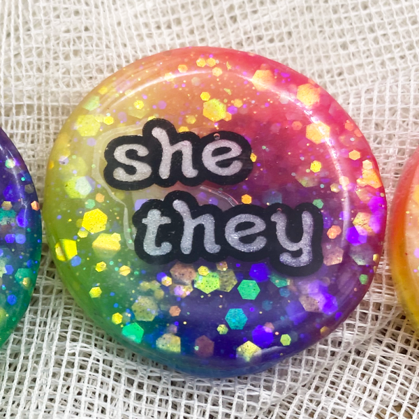 Glow in the dark rainbow pronoun badge with she/they pronoun label. Handmade in Melbourne by iridian.design.