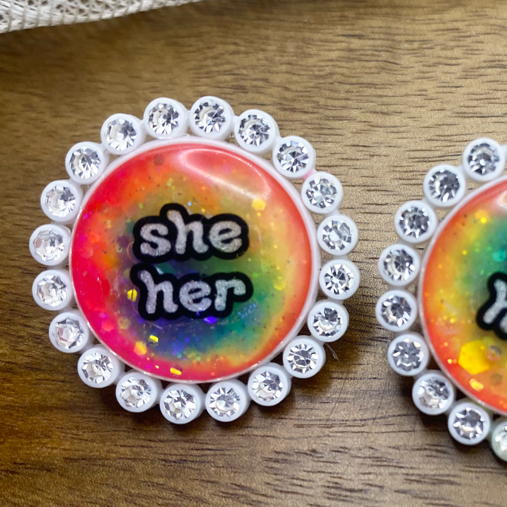 White rhinestone glowing rainbow pronoun pins with she/her label. Handmade in Melbourne by iridian.design.